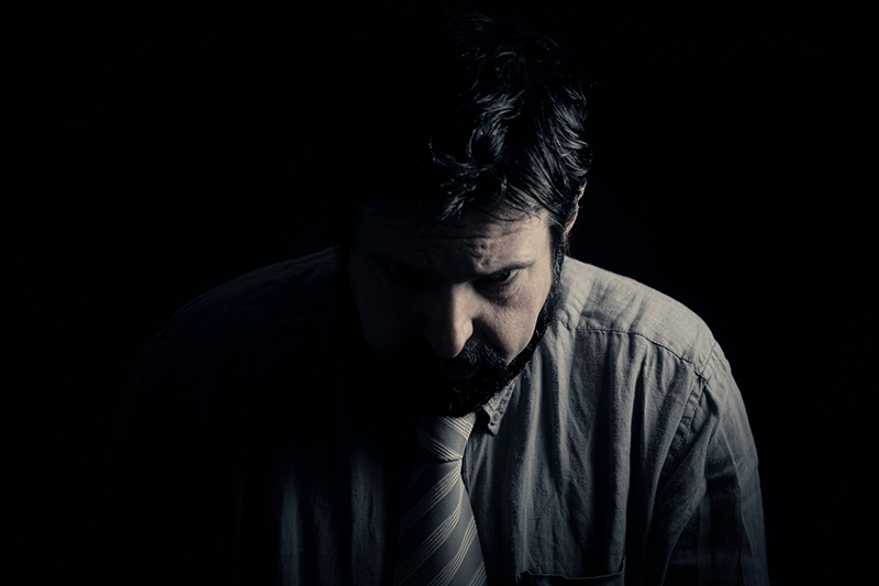 Photograph of a man looking down as though depressed, with a dark background and he is in shadow