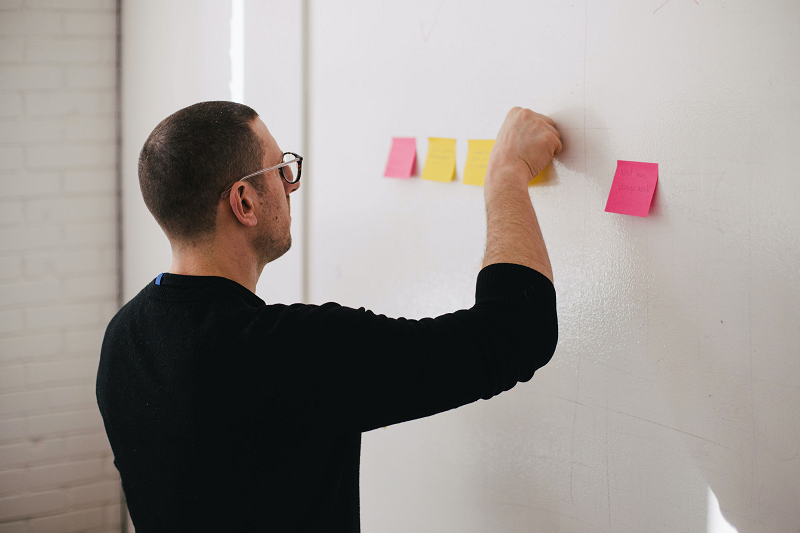 Man putting a coloured post-it note on a wall that has some on it already.